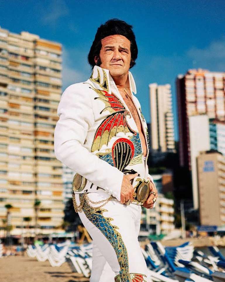 Full shot of adult male Elvis impersonator in patterned white jumpsuit standing on beach with buildings in background on clear, sunny day.