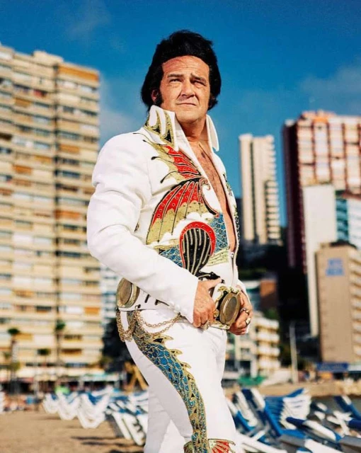 Full shot of adult male Elvis impersonator in patterned white jumpsuit standing on beach with buildings in background on clear, sunny day.