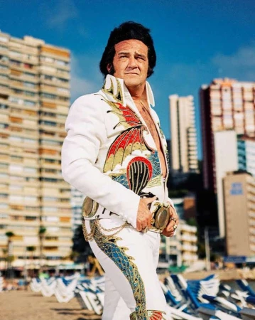 Full shot of adult male Elvis impersonator in patterned white jumpsuit standing on beach with buildings in background on clear, sunny day.