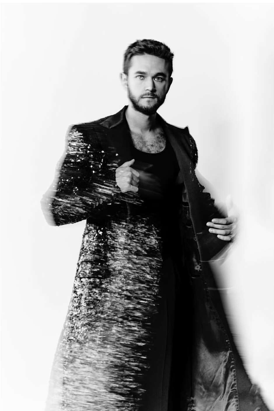 Black-and-white portrait of an adult male wearing a sequined coat and a tank top against a blurred white backdrop in a photo studio.