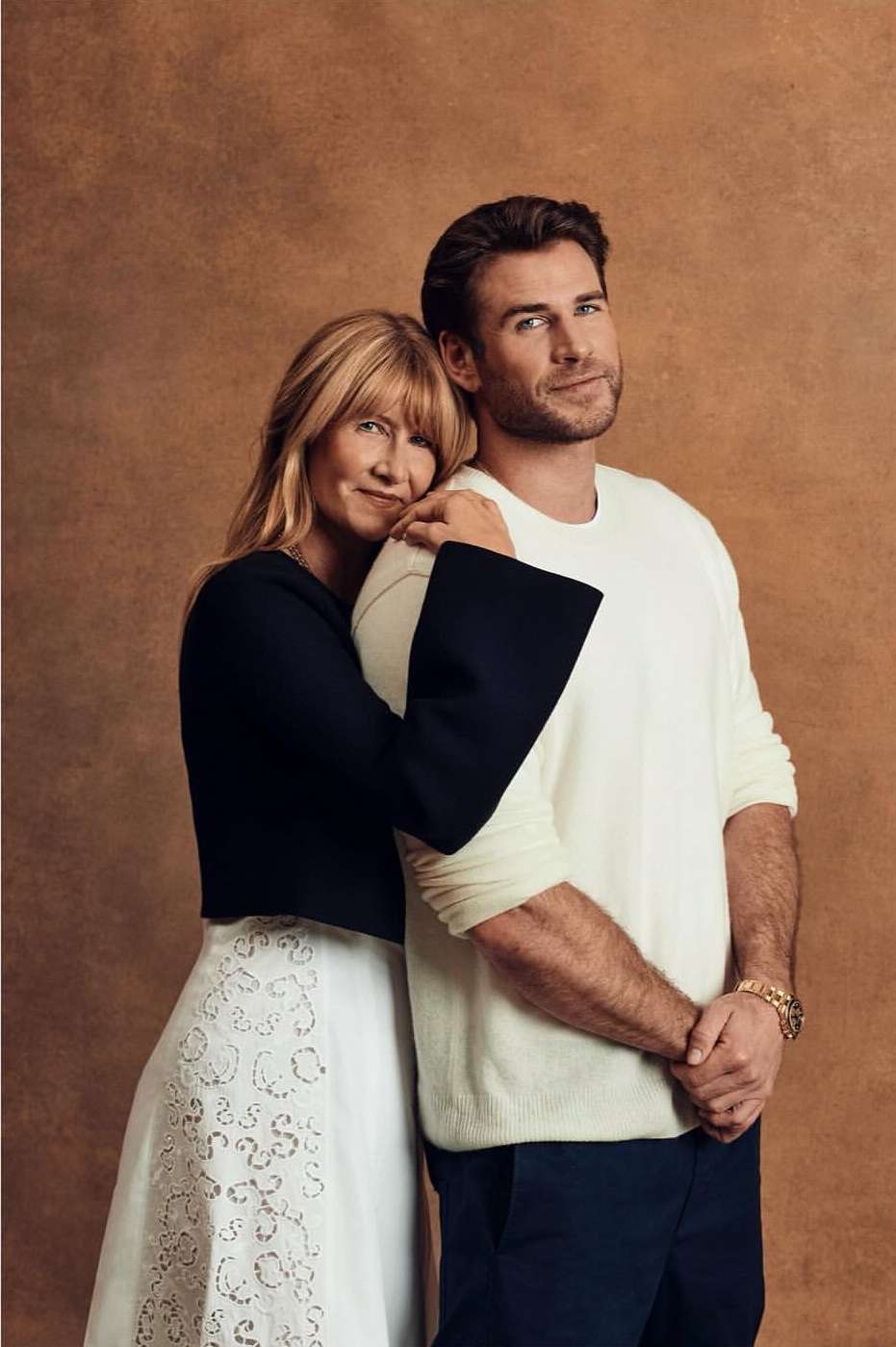 Portrait of adult male and adult female, arm around shoulder, wearing formal wear against a solid brown backdrop in a photo studio.