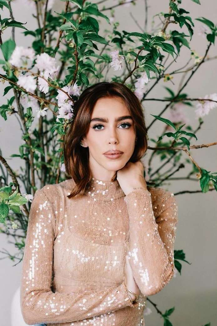Close-up portrait of a young adult woman with auburn hair wearing a sequined top, posing in front of a floral arrangement indoors.