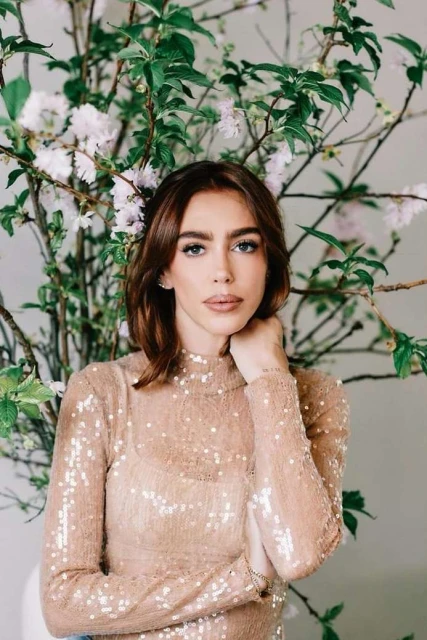 Close-up portrait of a young adult woman with auburn hair wearing a sequined top, posing in front of a floral arrangement indoors.