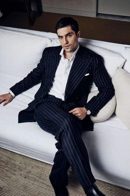 Well-dressed adult man in pinstripe suit and white shirt sits on a white sofa indoors with soft lighting, looking directly at the camera.