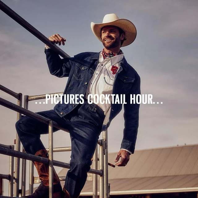 Man in a cowboy hat, denim jacket and jeans leans on a metal railing with a rural landscape in the background on a sunny day.