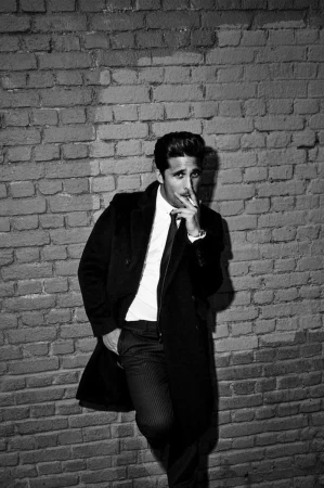 Black and white portrait of a man in a dark coat and tie leaning against a brick wall, smoking a cigarette.
