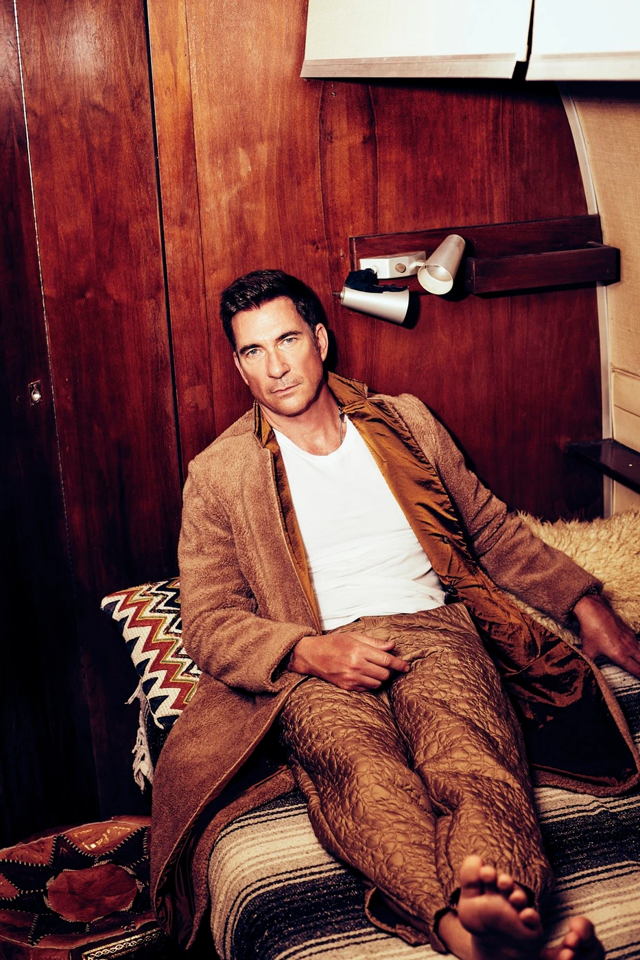 Man seated on a bed wearing a brown coat and trousers with a white t shirt against a wooden wall