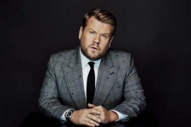 James Corden wearing a grey plaid suit with black star accents and a dark tie, photographed against a dark background.