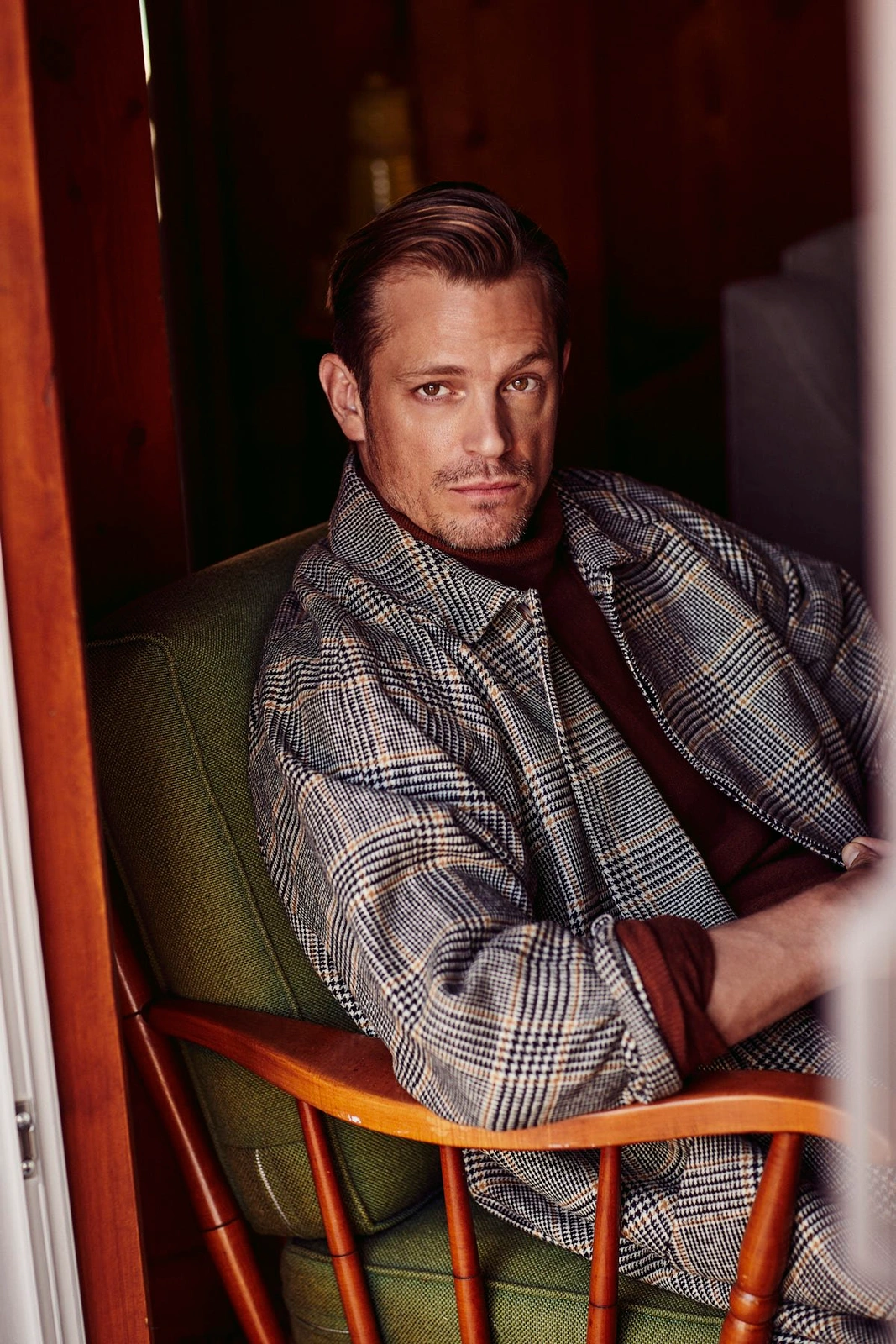 Man wearing plaid jacket and dark turtleneck seated in an armchair captured by Leigh Keily