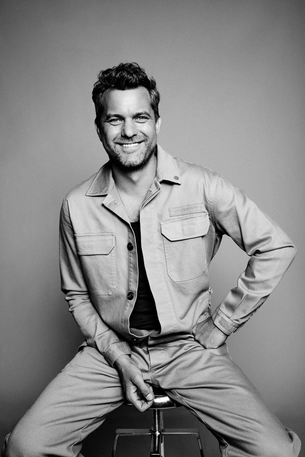 Portrait of actor Joshua Jackson smiling while seated on a stool wearing a utility jacket and matching trousers in a studio
