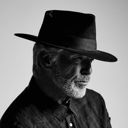 Monochrome portrait of an older man with a beard wearing a fedora and dark button-down shirt against a light background.