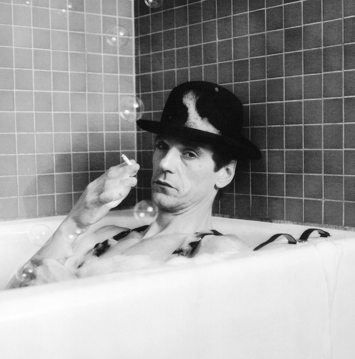 Monochrome portrait of a person in a bathtub with bubbles, wearing a hat and suspenders, holding a cigarette, with a tiled wall.