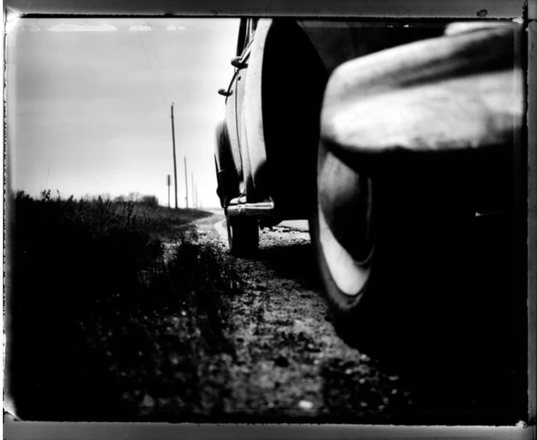 Monochrome image of a vintage car parked on a dirt road, captured from a low angle, with tall grass and utility poles in the background.