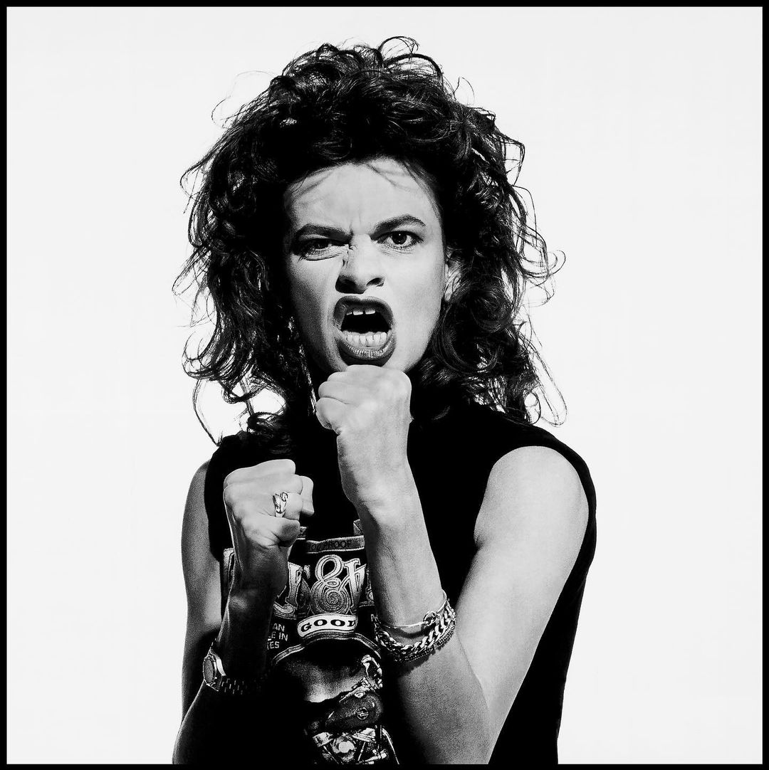 Monochrome portrait of a determined woman with curly hair and jewelry, raising her fists in a fighting pose against a white backdrop.