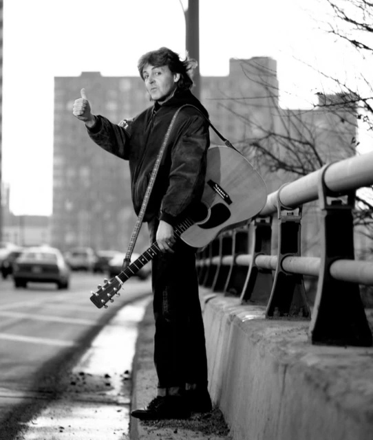Black and white shot of a man with a guitar giving a thumbs up on a city street, with cars and buildings in the background.