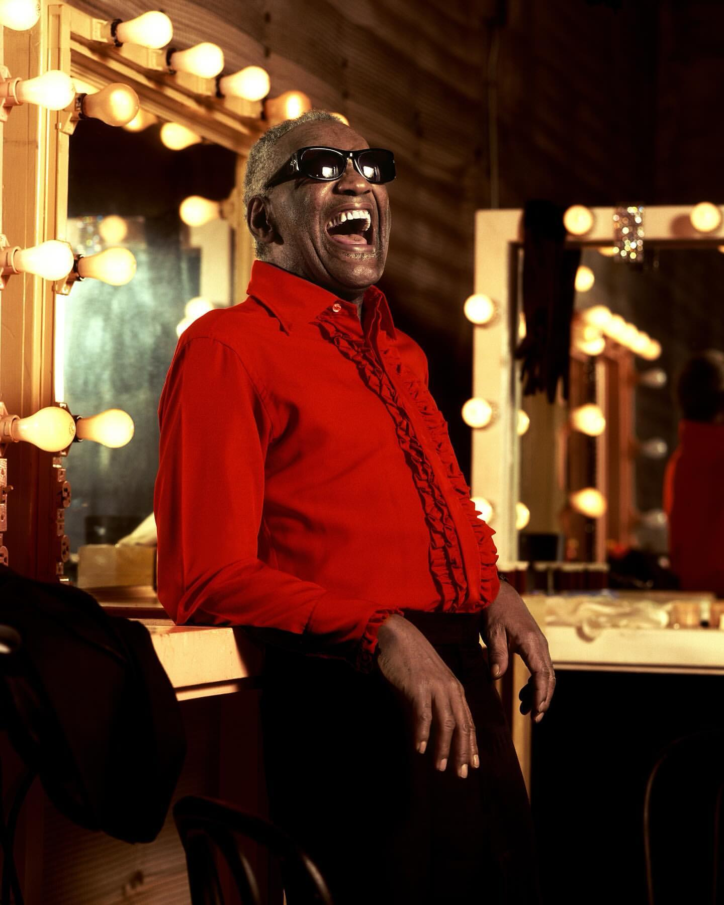 Ray Charles, an African-American man with dark glasses, smiles in a dressing room with lights and mirrors, wearing a red shirt and black pants.