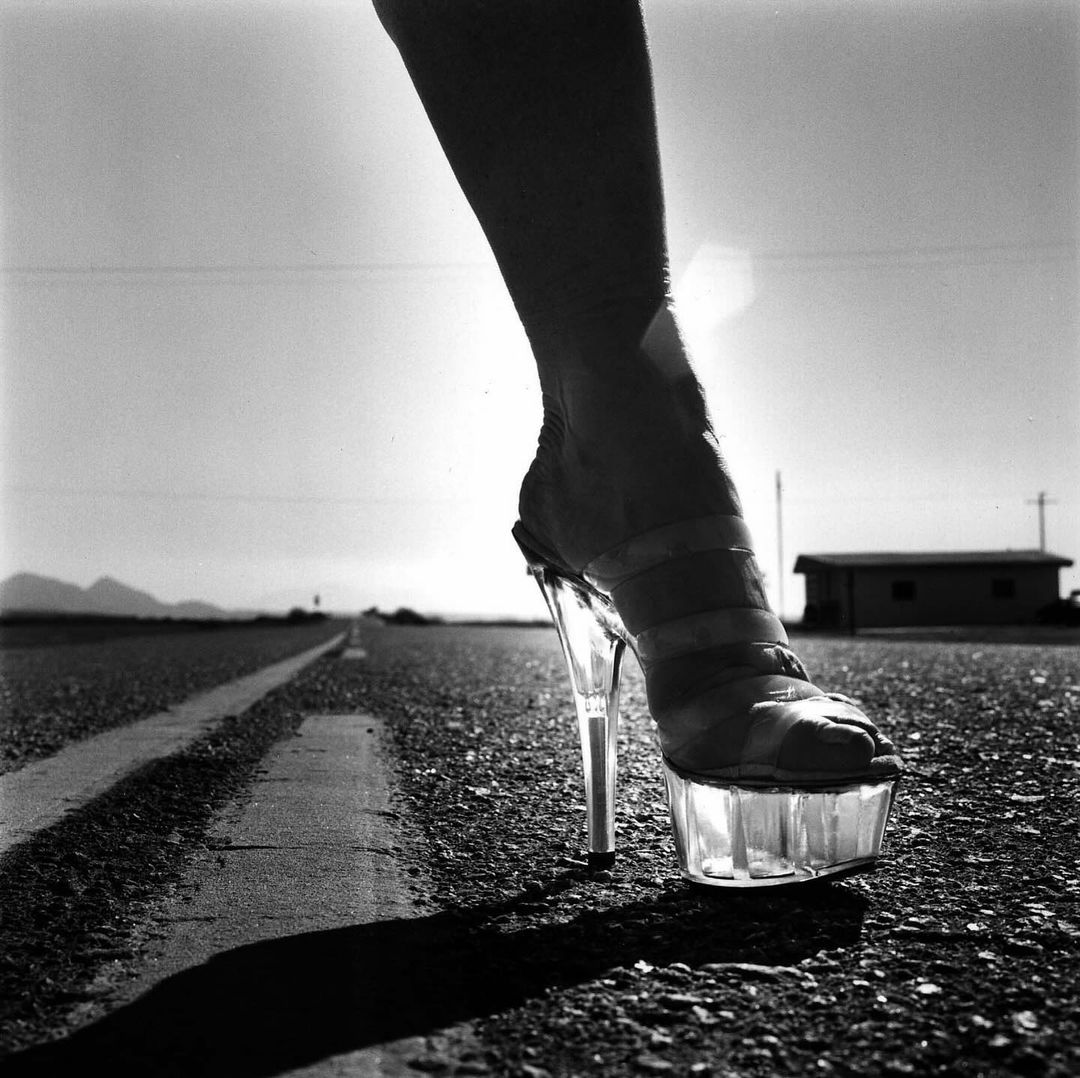 Striking monochrome image of a woman's foot in a clear platform high heel on a desolate road leading to a distant horizon.