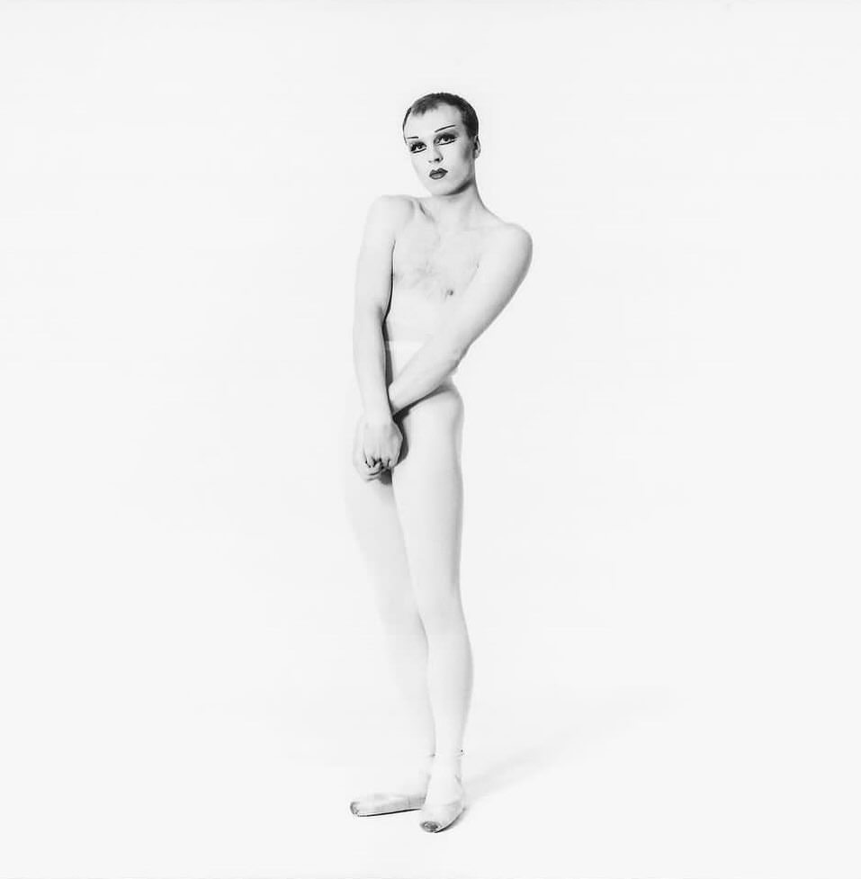 Adult male stands in a white studio wearing white tights and ballet shoes. The photograph is in black and white and displays modern dance.