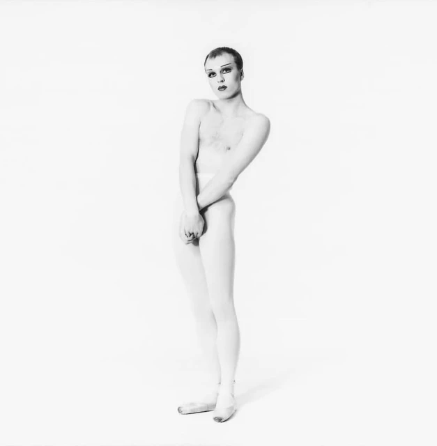 Adult male stands in a white studio wearing white tights and ballet shoes. The photograph is in black and white and displays modern dance.
