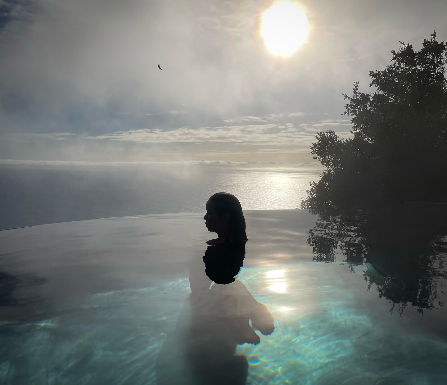 Person relaxes in an infinity pool overlooking ocean, sun, sky, and distant trees, creating a serene and scenic retreat.