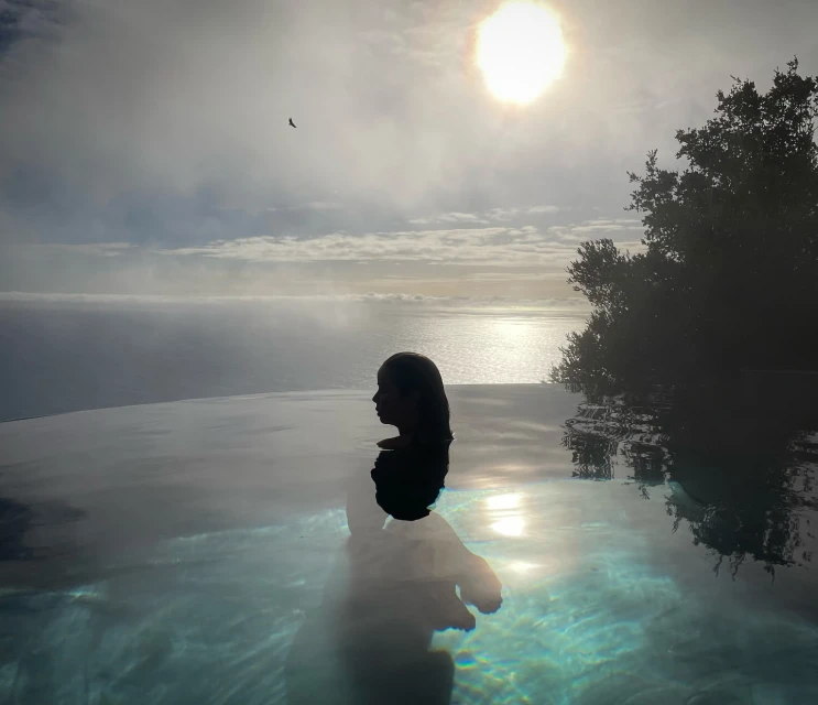 Person relaxes in an infinity pool overlooking ocean, sun, sky, and distant trees, creating a serene and scenic retreat.