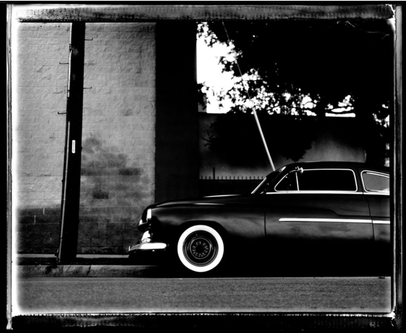 Black and white photograph of a vintage car parked on a city street, emphasizing its classic design and monochrome aesthetic, with tree shadows.