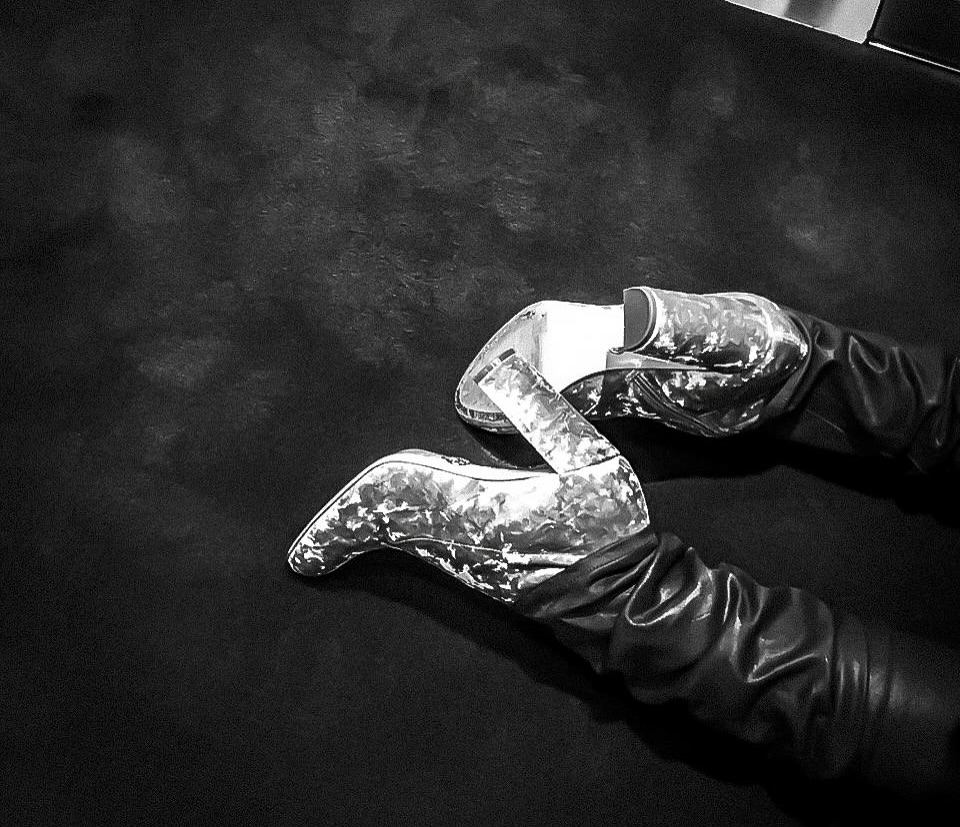 Close-up black and white shot showcasing a pair of silver high heels with patterned surfaces, worn with black leather opera gloves.