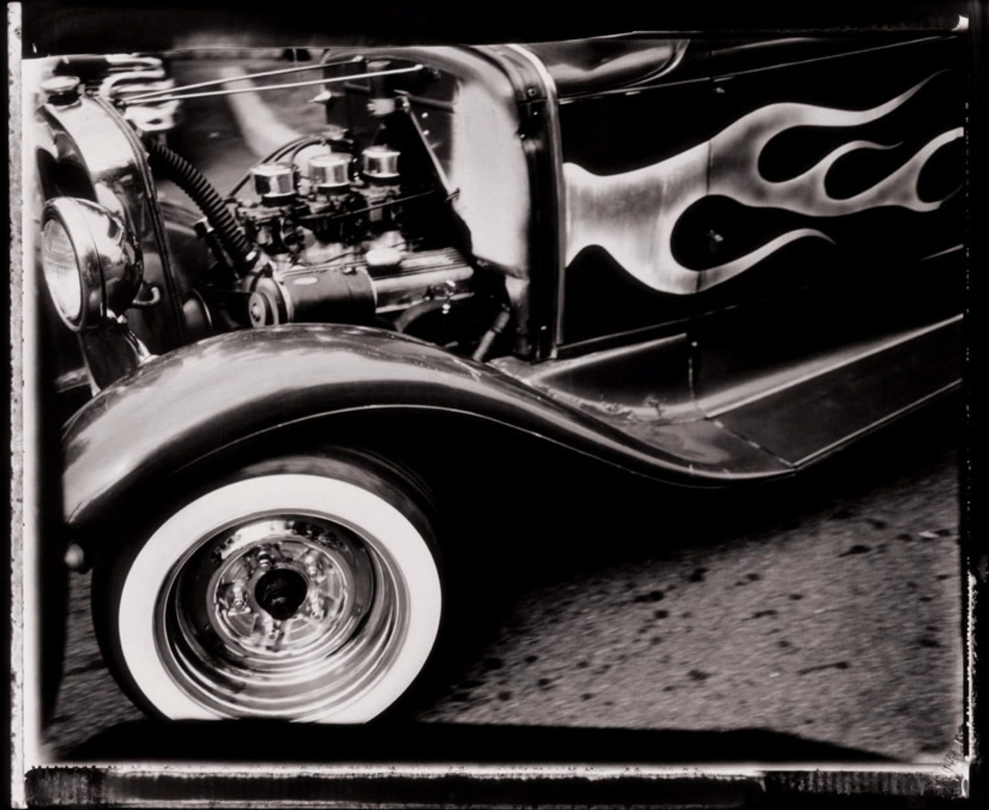 Black and white photograph of a vintage car with flame decals, showcasing the engine, whitewall tire, and detailed wheel.