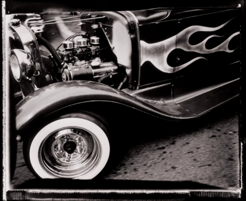 Black and white photograph of a vintage car with flame decals, showcasing the engine, whitewall tire, and detailed wheel.