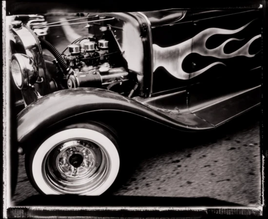 Black and white photograph of a vintage car with flame decals, showcasing the engine, whitewall tire, and detailed wheel.