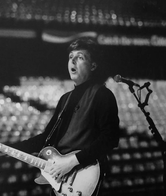 Black and white concert shot of Paul McCartney singing and playing guitar on stage with a microphone and blurred crowd in background.