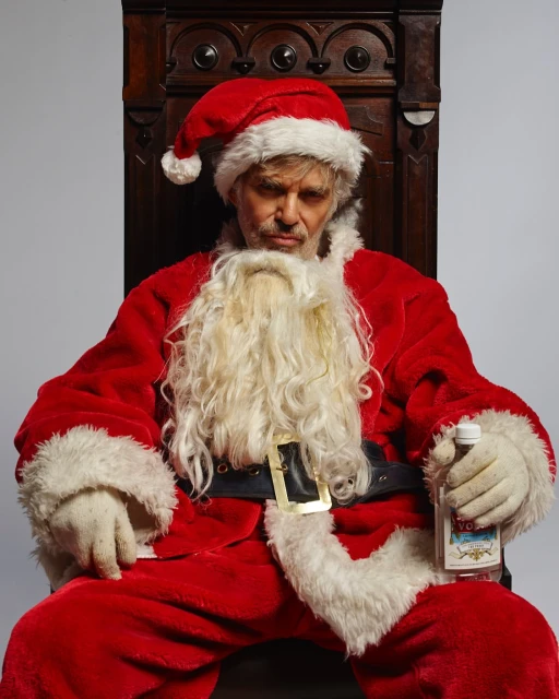Man in a Santa Claus costume sits on a wooden chair, holding a bottle of vodka against a grey backdrop in a studio setting.