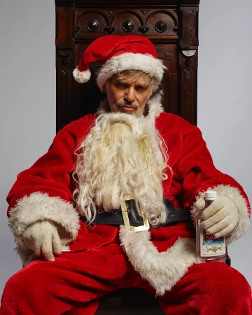 Man in a Santa Claus costume sits on a wooden chair, holding a bottle of vodka against a grey backdrop in a studio setting.