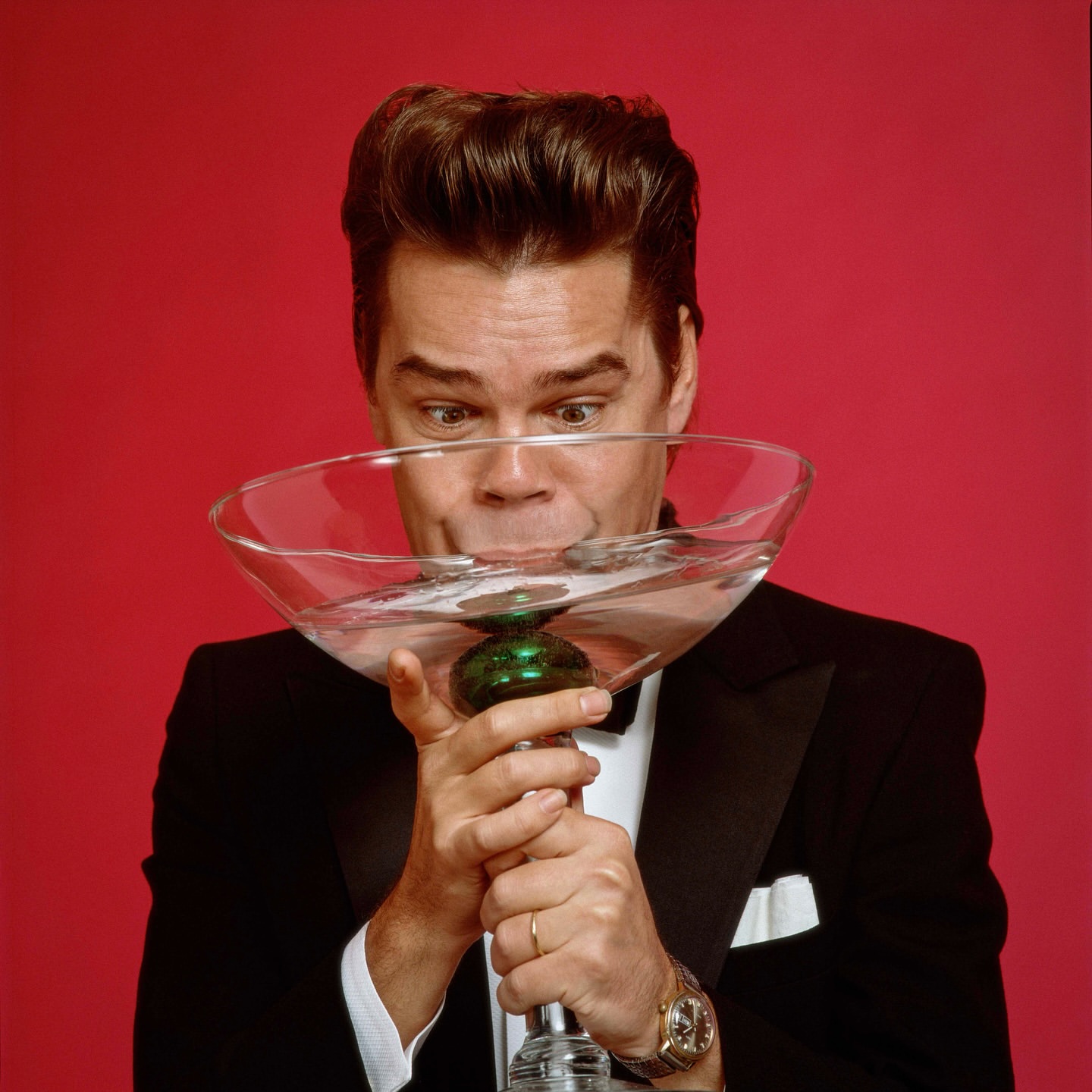 Comedian and actor Will Forte in tuxedo drinks from a large novelty martini glass in a studio shot against a red backdrop.