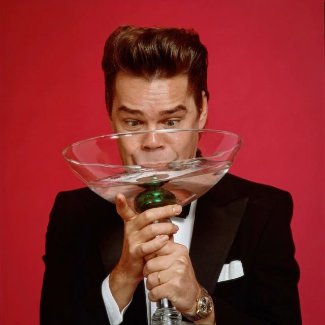 Comedian and actor Will Forte in tuxedo drinks from a large novelty martini glass in a studio shot against a red backdrop.