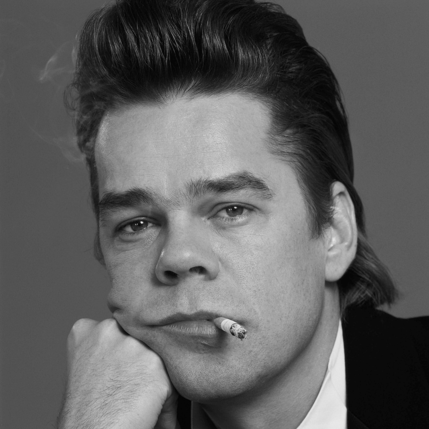 Black and white portrait of an adult male smoking a cigarette, resting his head on his hand, with a plain grey background.