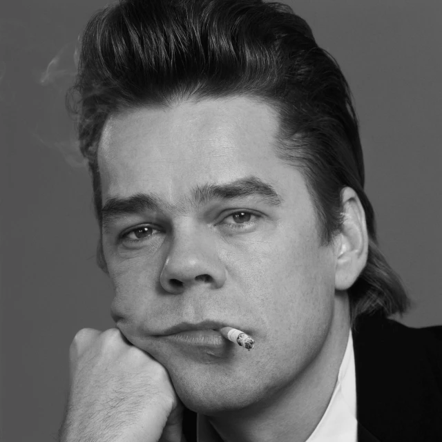 Black and white portrait of an adult male smoking a cigarette, resting his head on his hand, with a plain grey background.