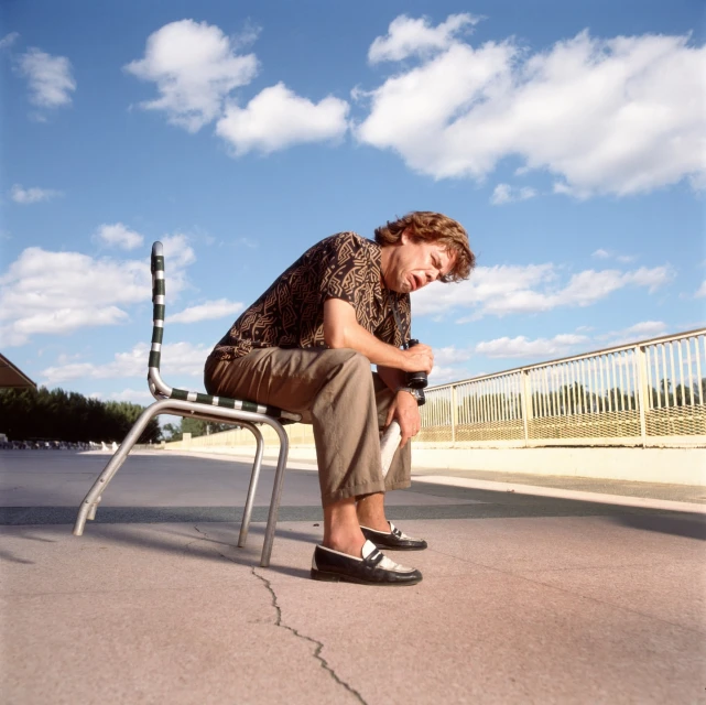 Person sits on a chair outside with a drink on a sunny day under blue sky with clouds, wearing a patterned shirt and brown pants.