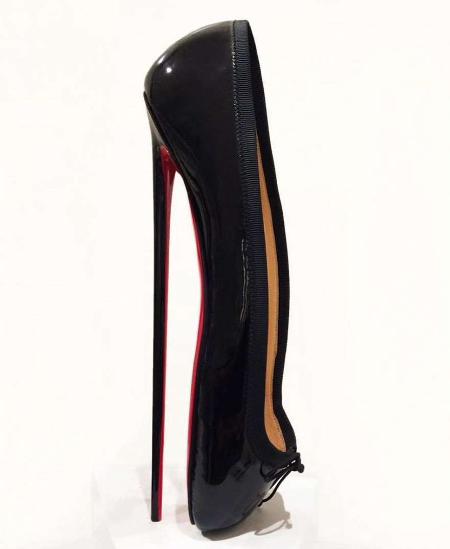 A black patent leather ballet flat with a stiletto heel and red sole is isolated on a white background in a studio shot.
