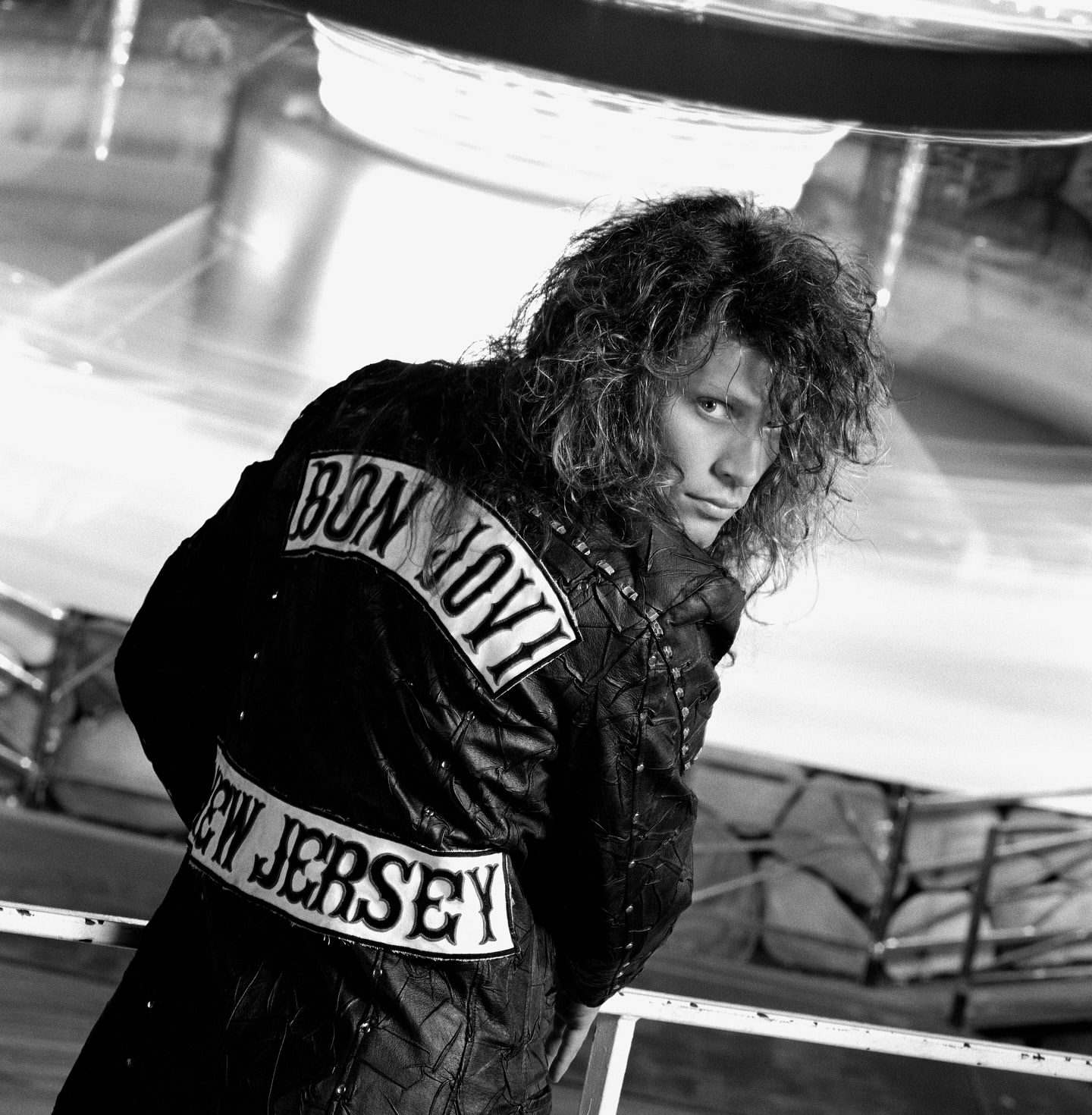 Black and white portrait of a man with long curly hair wearing a Bon Jovi 'New Jersey' jacket looking back at the camera.