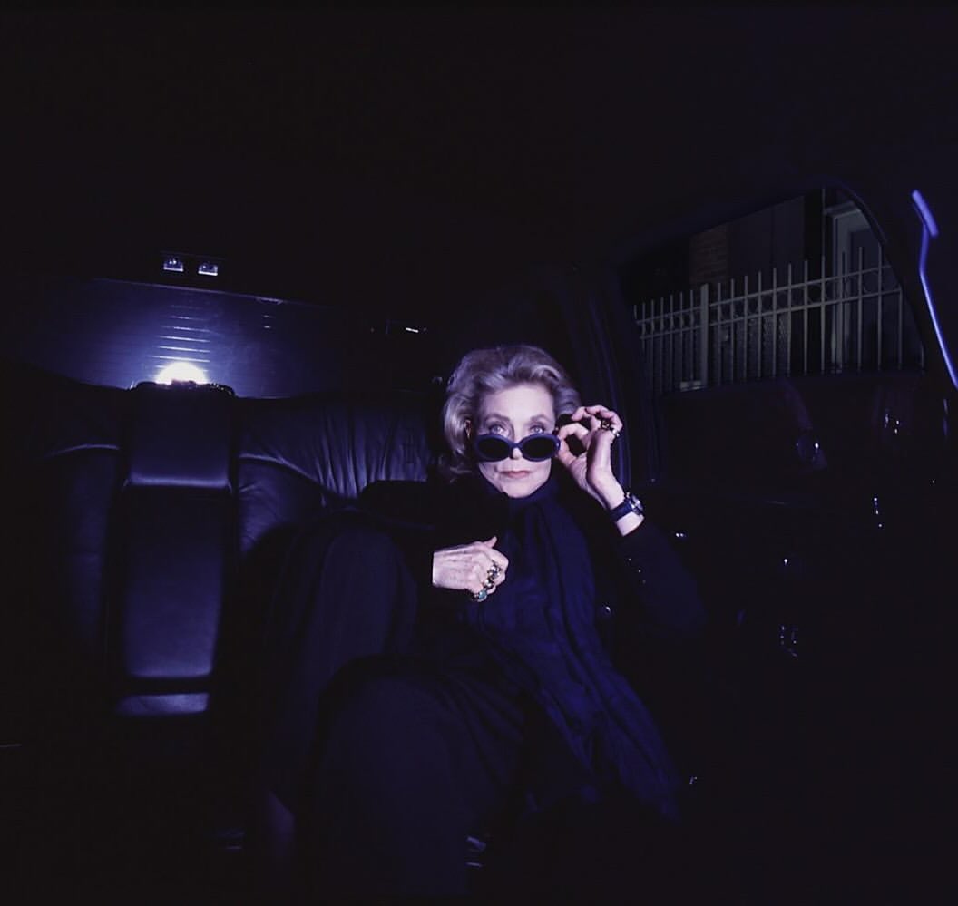 Mature woman in sunglasses sitting in the back seat of a car at night, illuminated by interior lights, wearing a dark coat and scarf.