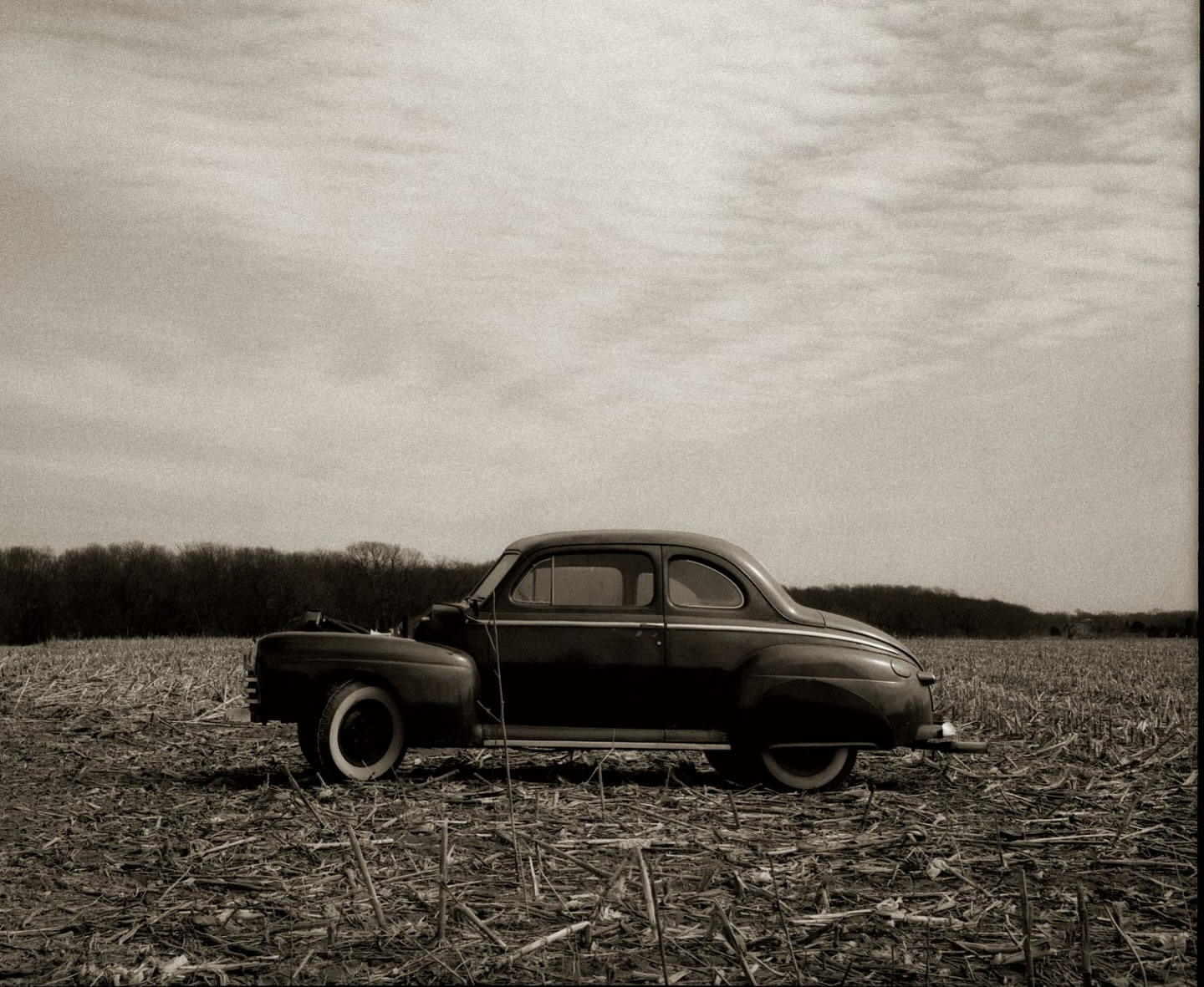 Vintage black car parked in a harvested cornfield under a cloudy sky, evoking a sense of rural nostalgia and classic automotive design.