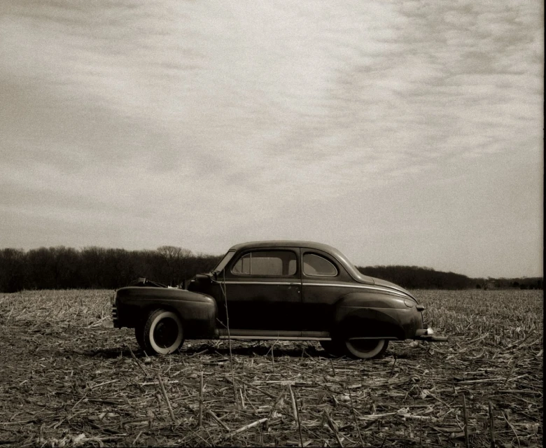 Vintage black car parked in a harvested cornfield under a cloudy sky, evoking a sense of rural nostalgia and classic automotive design.