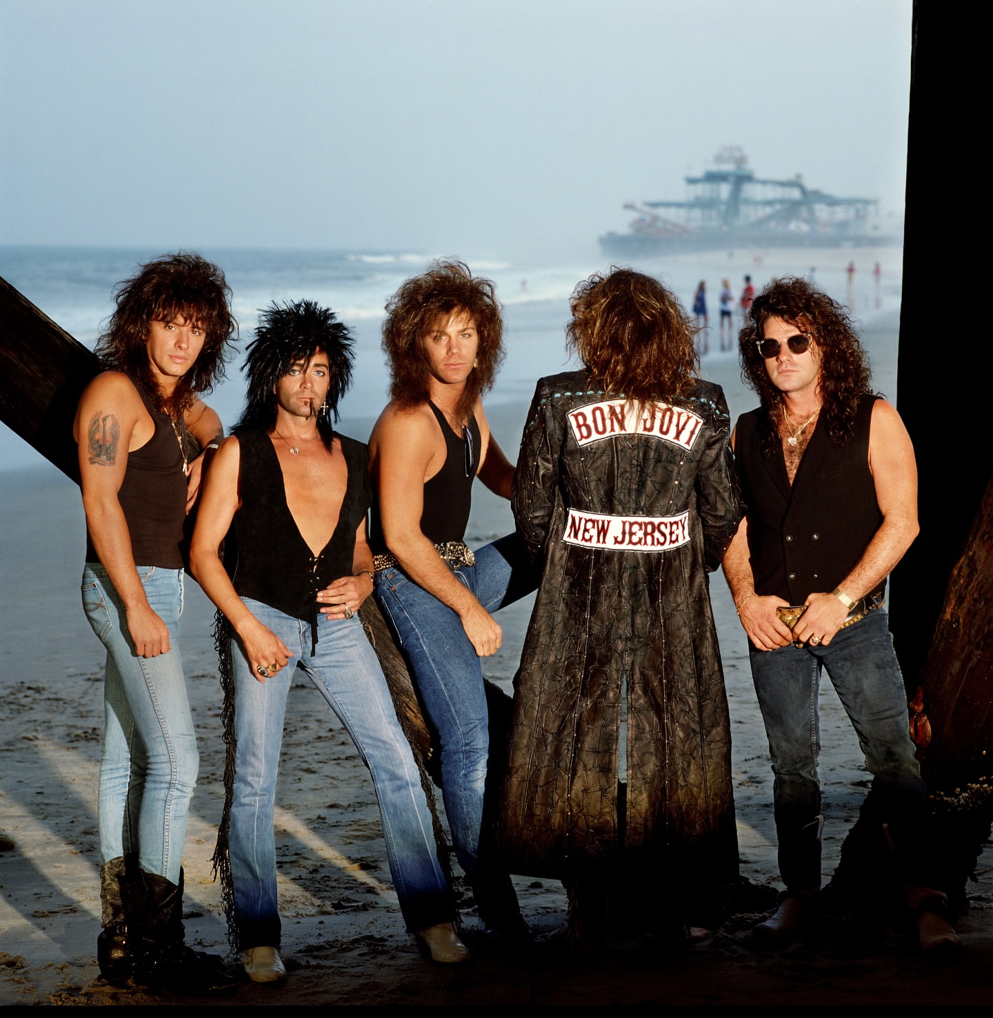 Five members of Bon Jovi pose on a sandy beach, one wearing a leather jacket with the band's name, ocean and pier in background.