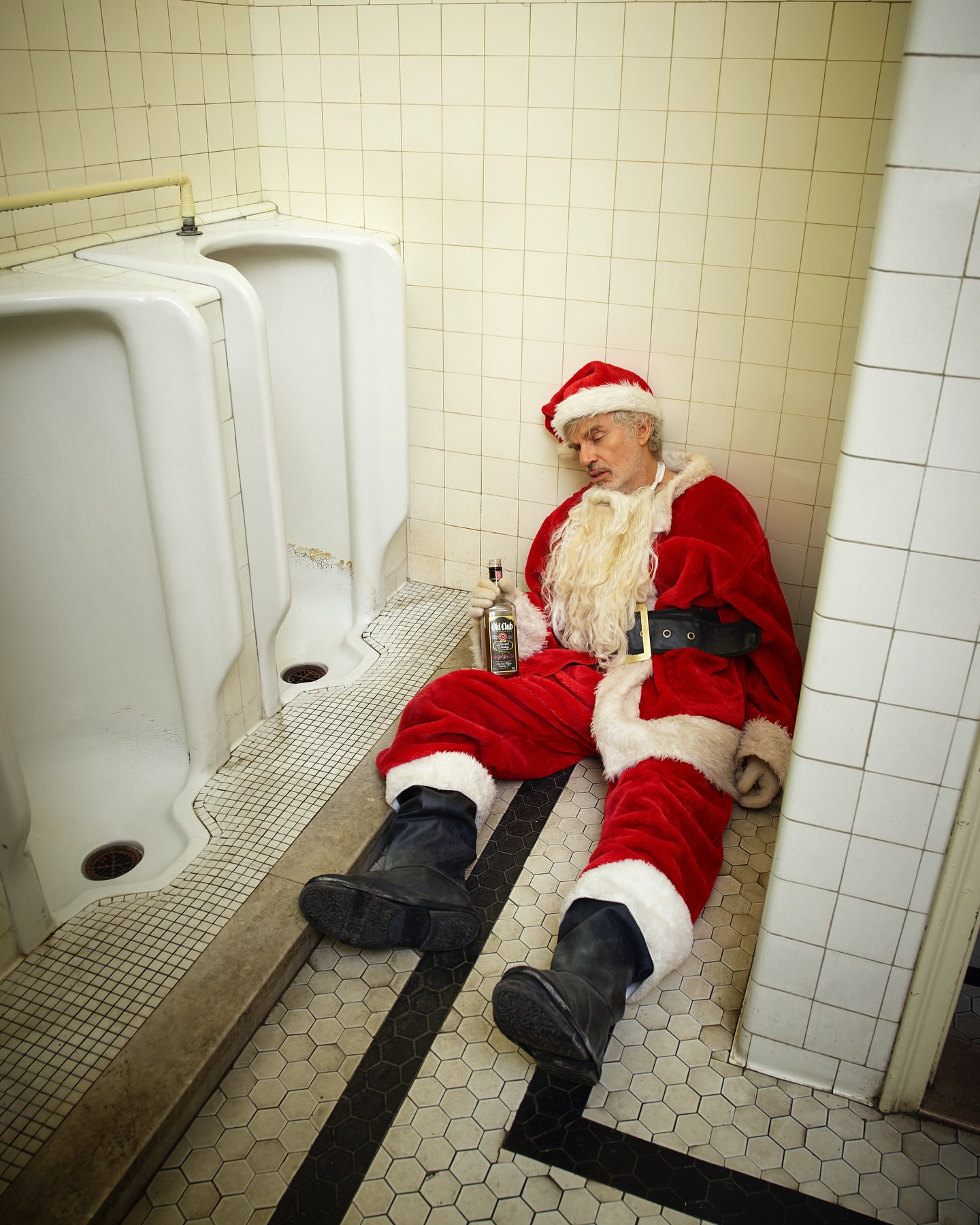 Adult male dressed as Santa Claus sits on the floor of a public bathroom next to white urinals with a bottle of alcohol.