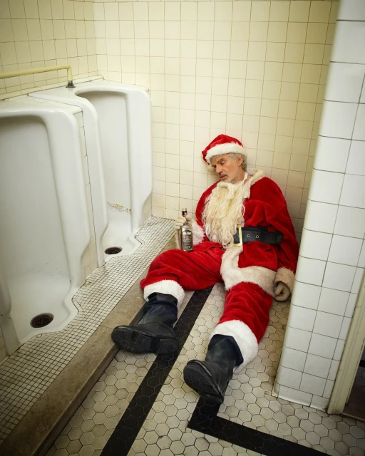 Adult male dressed as Santa Claus sits on the floor of a public bathroom next to white urinals with a bottle of alcohol.
