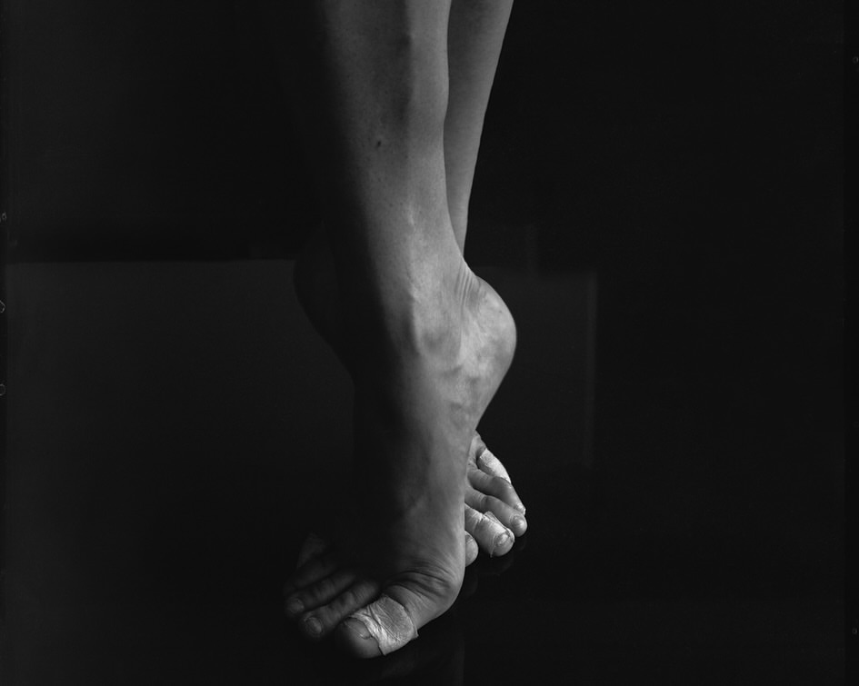 Black and white image of human legs and feet with crossed ankles, showing the human body on a reflective surface against a dark background.