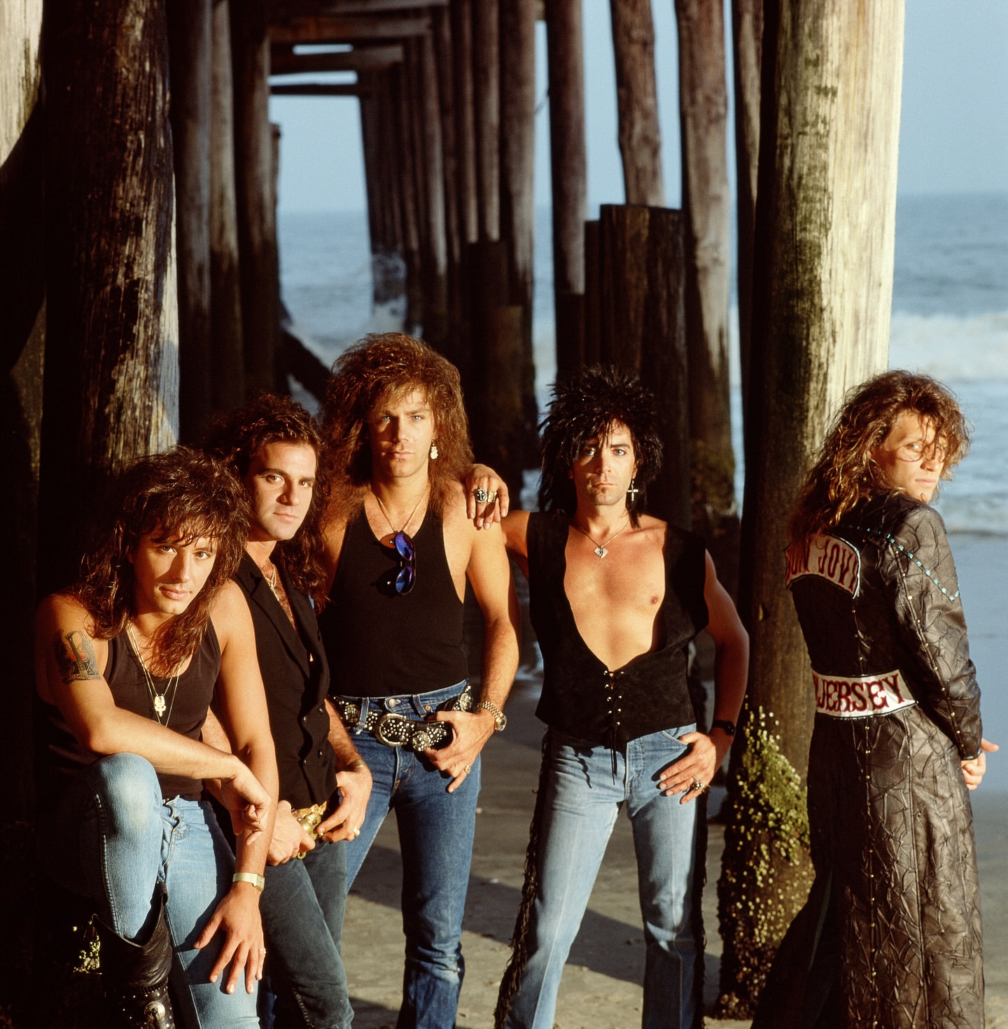 Glam metal band posing under a pier on a beach. Five adult males with long hair and makeup. Band members are wearing casual clothing and jewelry.