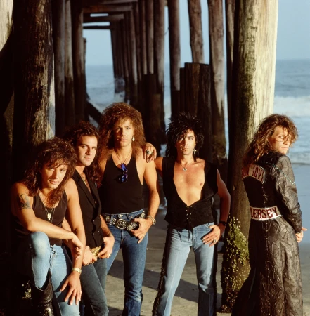 Glam metal band posing under a pier on a beach. Five adult males with long hair and makeup. Band members are wearing casual clothing and jewelry.