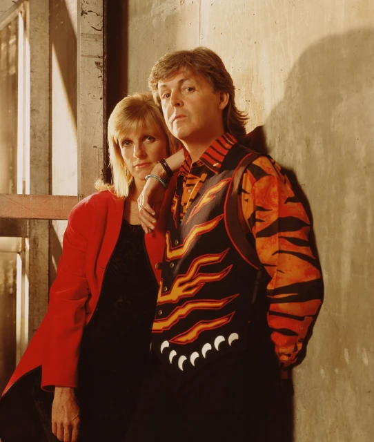 Portrait of adult male and female wearing 1980s fashion, posing against a concrete wall with metal framing in the background.